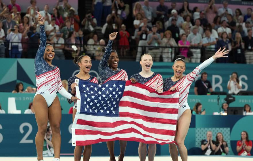 Five gymnasts from Team USA jump and scream while holding the American flag just after winning the 2024 Paris Olympics women's team competition