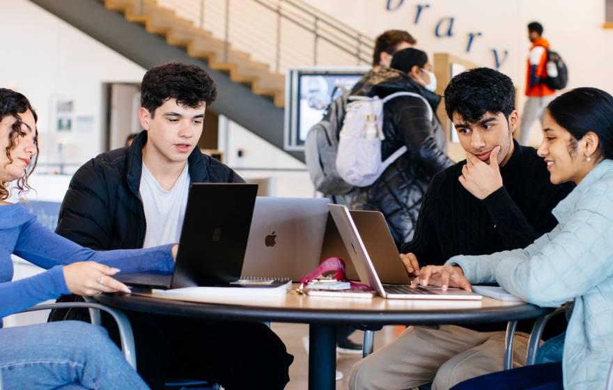 A group of students studying together at a table in a busy campus space, with a sign that says "Library" in the background. 
