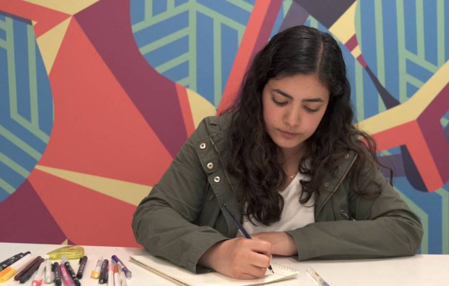 Frida writes in her notebook in front of a colorful wall