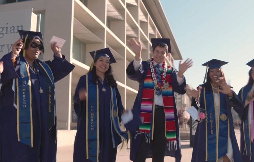 A row of students in graduate robes, caps and stoles wave hands excitedly in front of a library