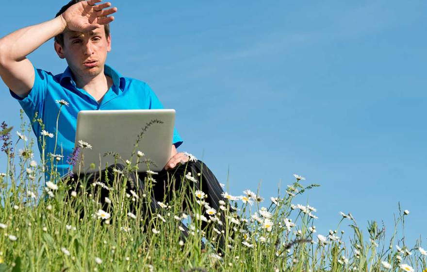 a man outside, sitting in a field with a laptop, wiping sweat off his brow