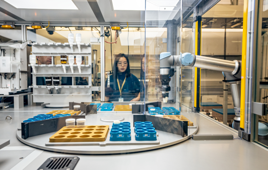 Young woman researcher looking through glass at a robotic arm preparing to handle samples