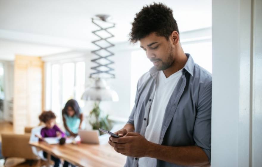 A man uses a smartphone while his kids are at a table in the background