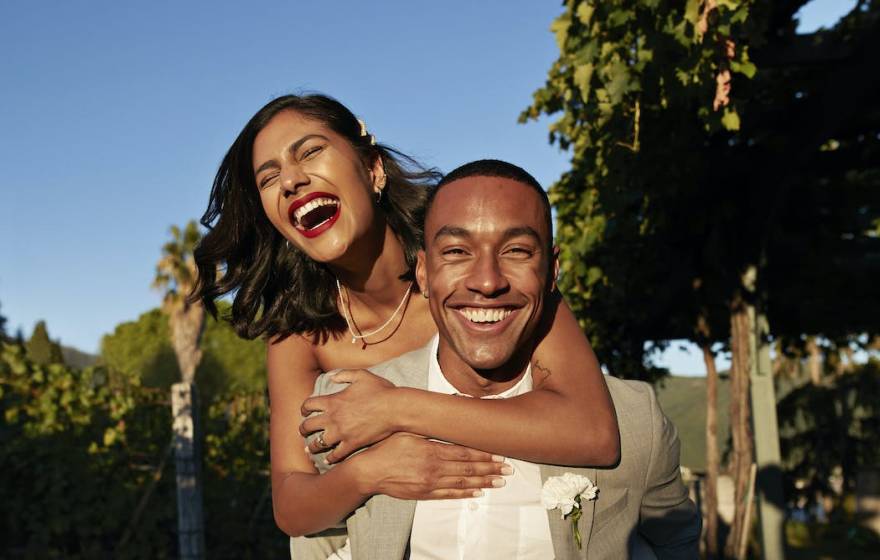 A happy young Latina woman laughing while hanging off the back of a happy young Black man, both in wedding outfits