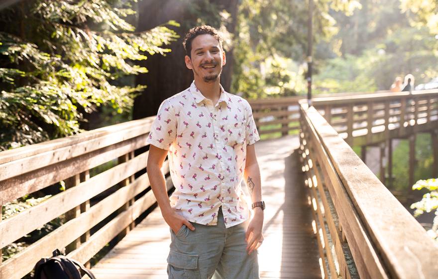 A smiling young man standing on a bridge surrounded by redwood trees