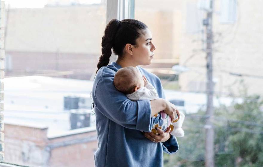 A woman with a long ponytail holds a baby and looks out the window with a trouble expression on her face