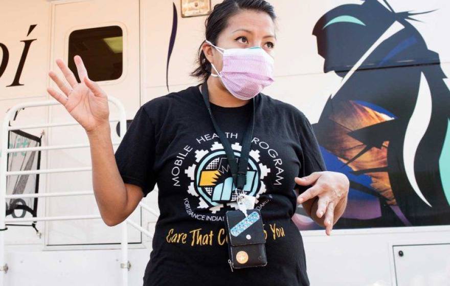 A young woman wearing a black t-shirt and a pink face mask gesticulates with her hands standing in front of a trailer