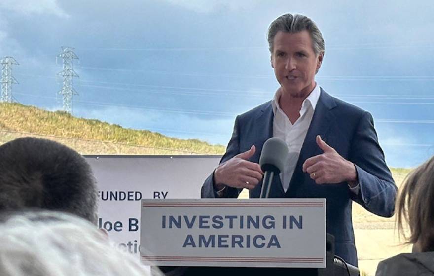 Governor Newsom speaking at a podium that says Investing in America, with countryside behind him