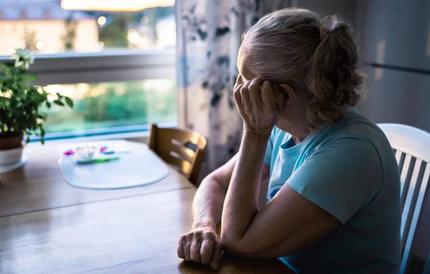 Older woman in blue shirt sitting at kitchen table, gazing pensively out window
