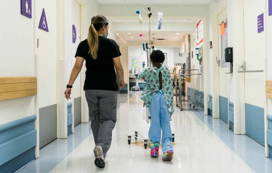 a nurse wearing a mask walks with a young Black patient around a hospital unit