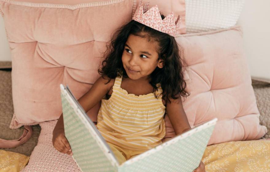 A young South Asian girl reads a book while wearing a crown and smiles