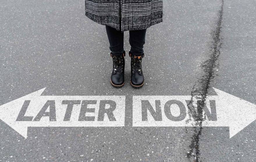 View of a person standing on pavement with only their legs and boots visible, positioned above two large white arrows painted on the ground—one pointing left labeled “LATER” and one pointing right labeled “NOW.”