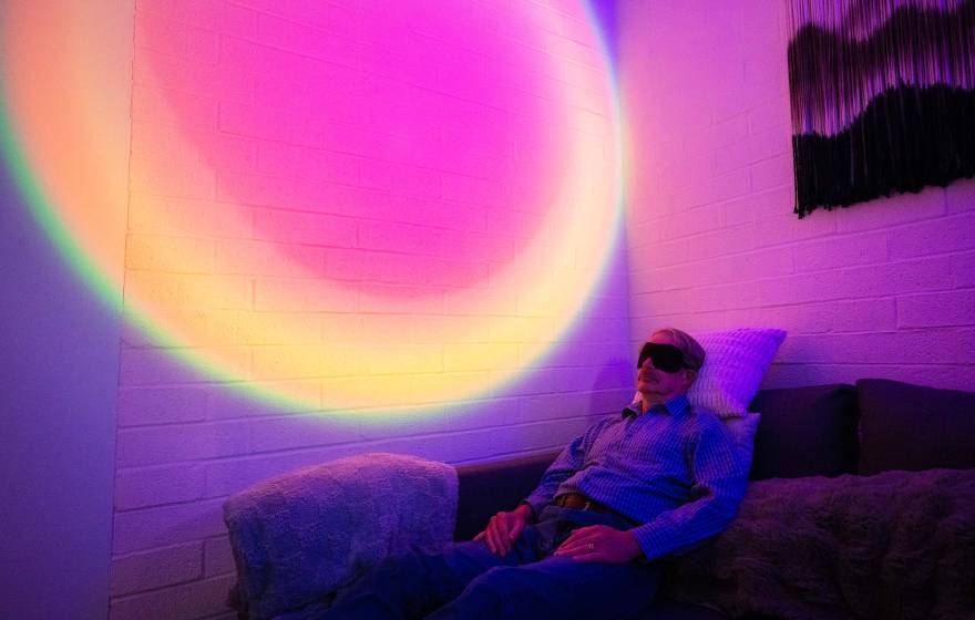 A man wearing an eye mask reclines on a couch in a darkened room. Behind him, the wall is illuminated with a circular, rainbow-colored light