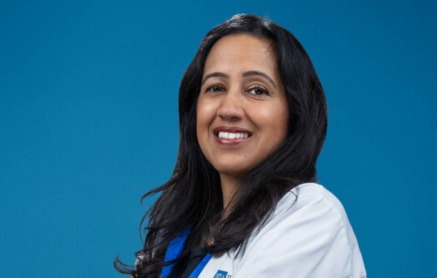 A portrait of a smiling woman with long hair in a white medical coat on a blue background