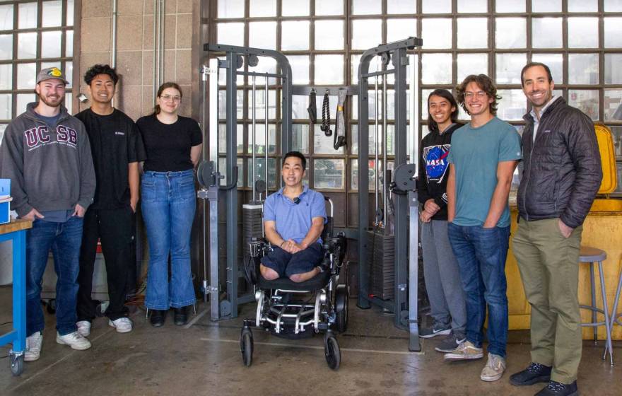 A team of students and a professor stands around a young man in a wheelchair underneath a workout machine