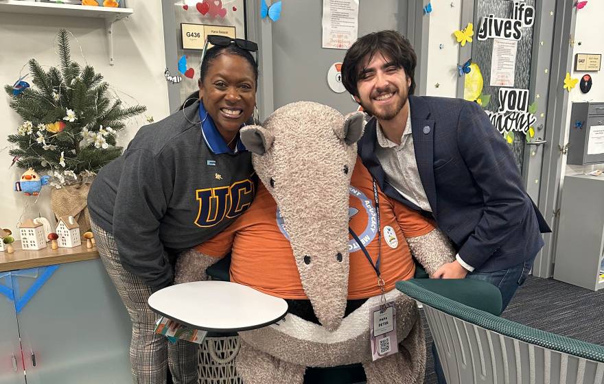 In a student center space, on the left, a woman with a sweatshirt that says UC and on the right, a young man with a beard smiling; center, a stuffed Anteater, the UC Irvine mascot