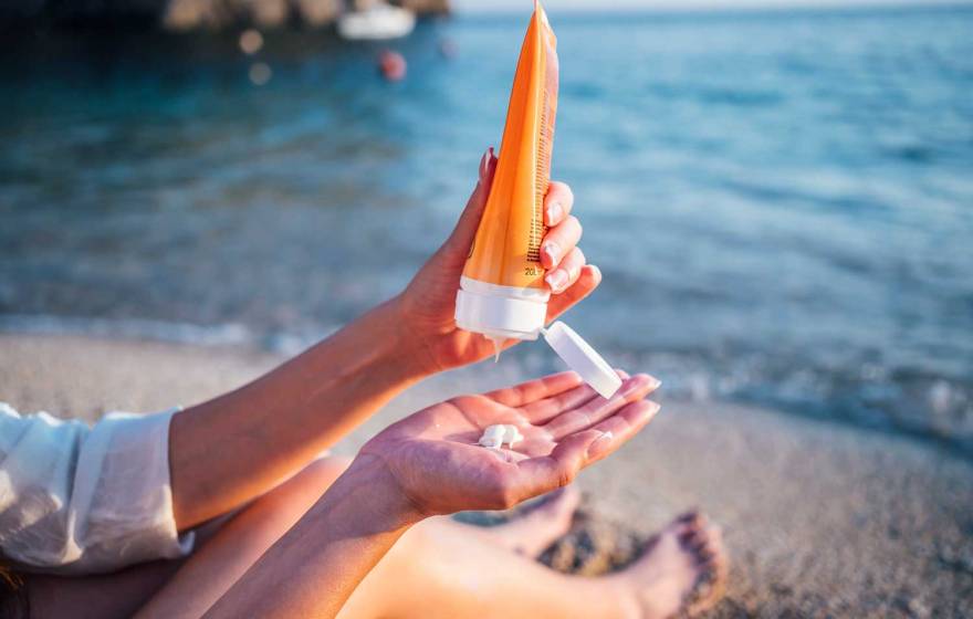 Someone sitting on the beach holds a tube of sunscreen in one hand with the other with a dab of sunscreen on it