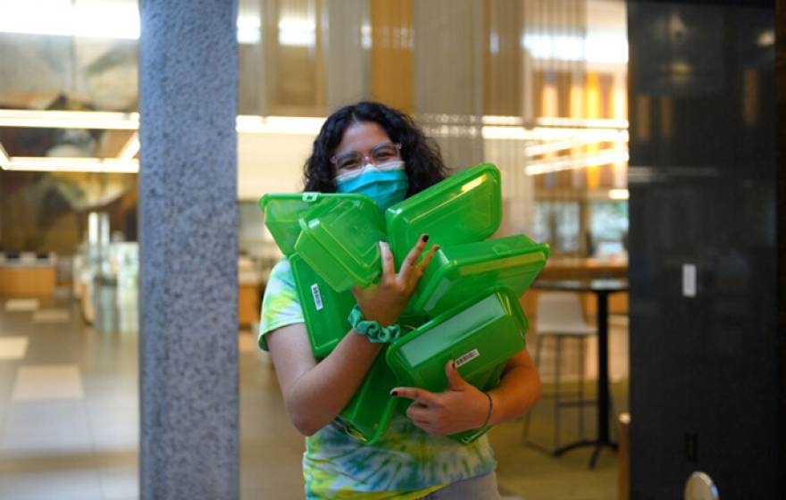 Woman in a mask carrying an armload of green reuseable food containers