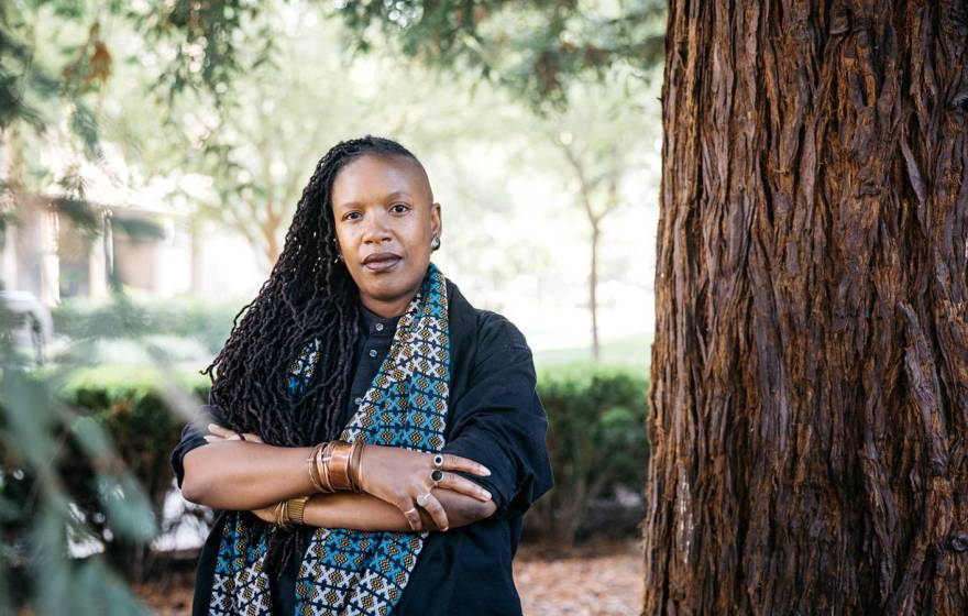 E. Tendayi Achiume, African American woman, poses outside, arms crossed, next to a redwood