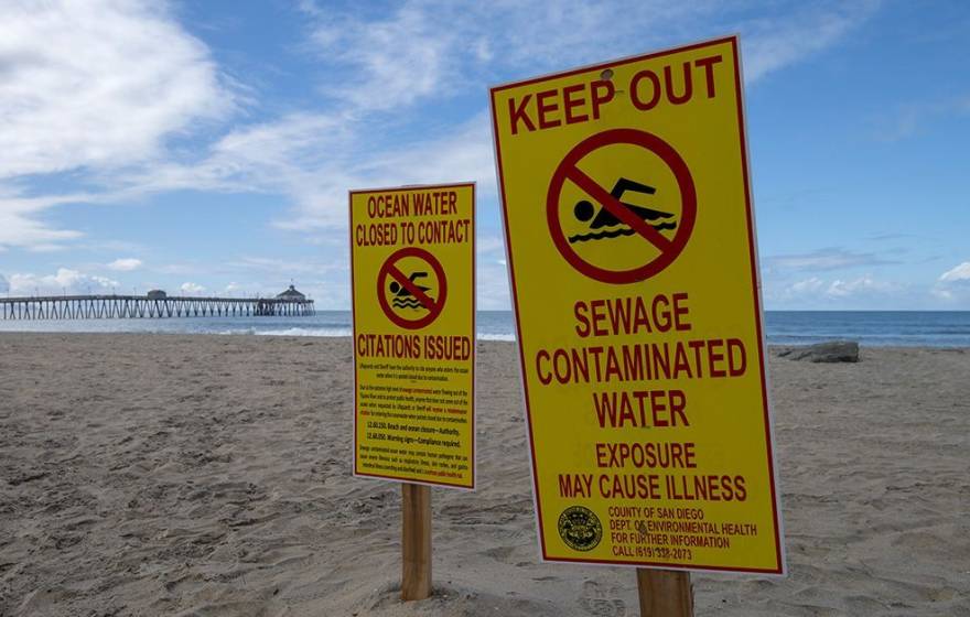 An empty beach with a pier in the background on a nice day bears signs that say Keep Out Sewage Contaminated Water and Ocean Water Closed to Contact Citations Issued