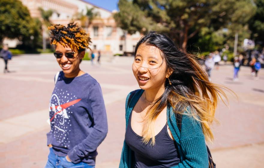 A young Black woman with a NASA sweatshirt and a young Asian woman with a green cardigan walk along a campus smiling