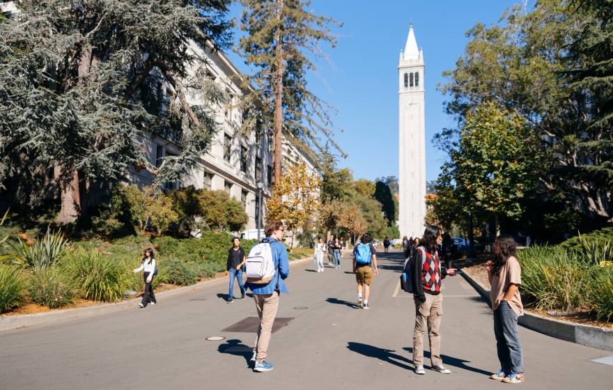 Students walk and talk on a tree-lined wide walkway in front of Sather Tower on the UC Berkeley campus