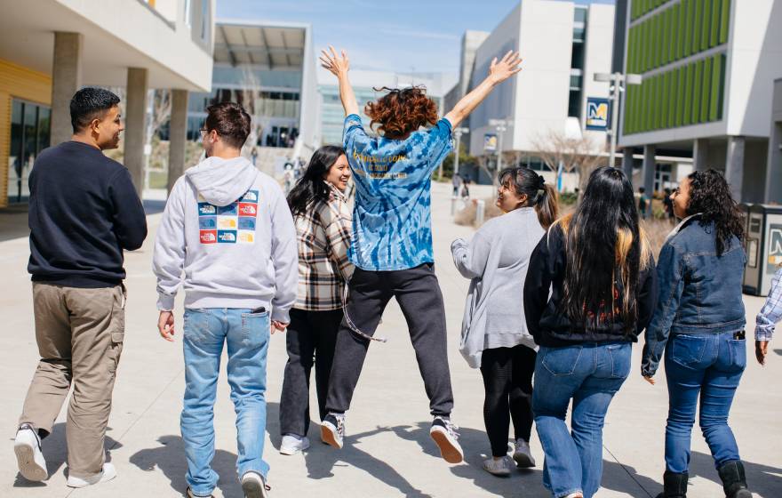 Viewed from behind, a diverse group of students walks along the UC Merced campus, the center student jumping exuberantly