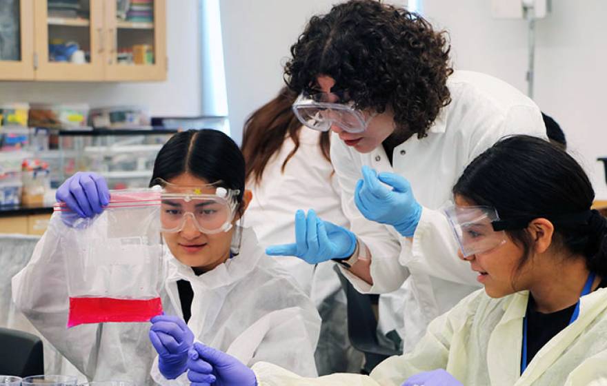 A college student in PPE shows two high schoolers in PPE how to collect a sample in a lab