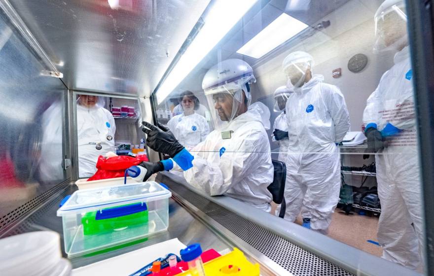 A group of students in PPE watch someone pour something behind protective glass