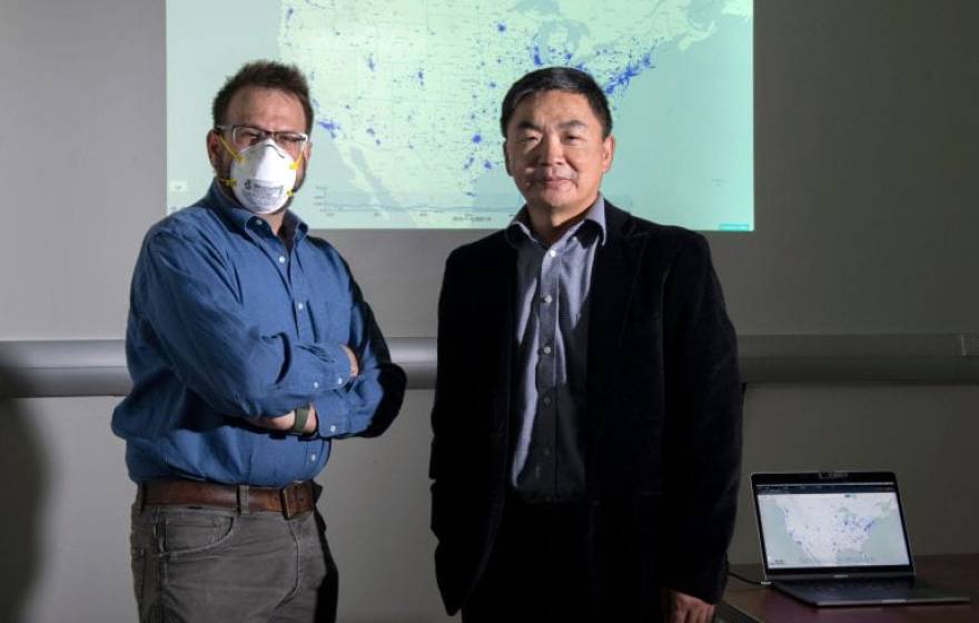 Two men standing together with a projector screen behind them, one wearing a face mask