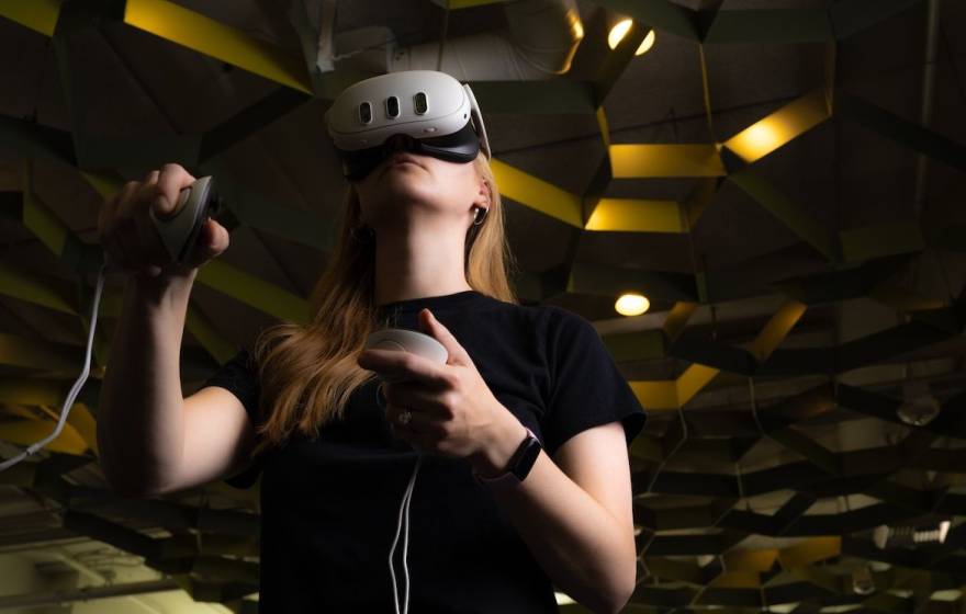 A student with a virtual reality headset and two controllers stands in a room with a honeycomb-esque roof
