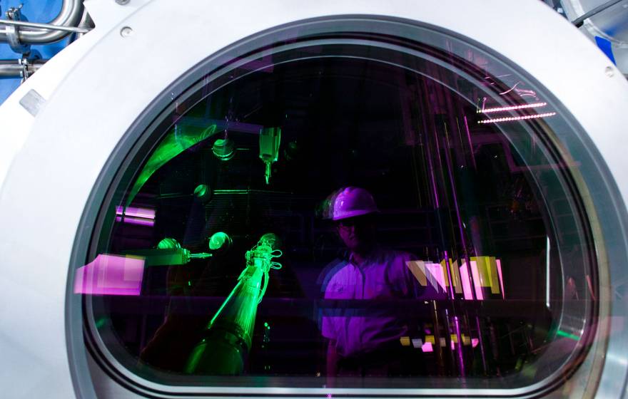 A person wearing a white hard hat is reflected in the circular viewing window of a large scientific instrument, while bright green laser beams and purple lighting illuminate the interior of a laboratory filled with optical equipment and metal piping.