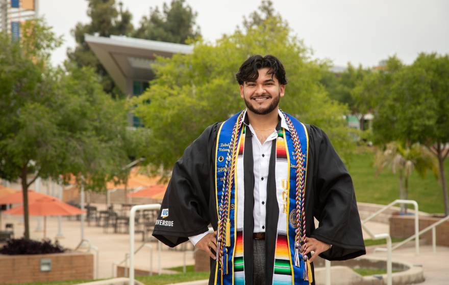 man with hands on hips wearing graduation gown and sashes on UC Riverside campus