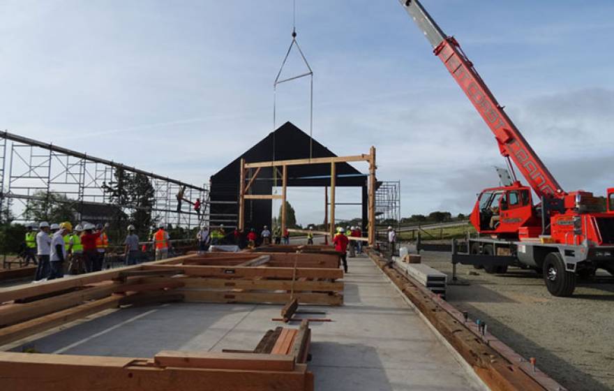 The first &quot;bent,&quot; a cross beam, posts, and supports joined by wooden pegs, is hoisted into place by a crane early Saturday morning while others, already assembled, await their turn on the new barn&#039;s concrete slab.