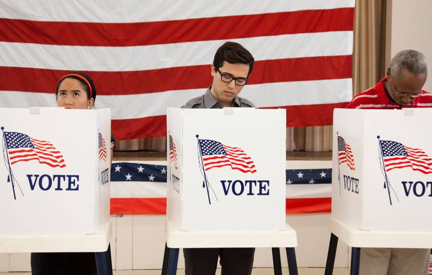 3 people stand at voting booths in front of a wall-sized American flag