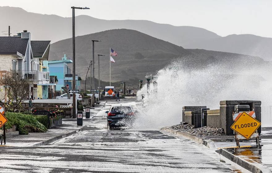 A wave crashes into a town street a blue car is driving on in Pacifica