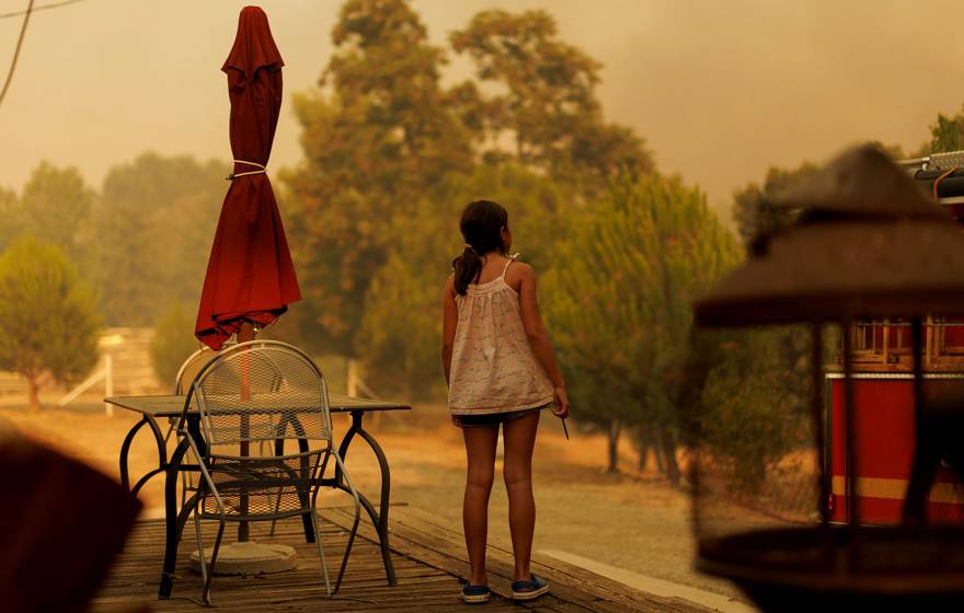 A school-aged girl photographed from behind, standing on the deck of her house, looking at a smoky sky