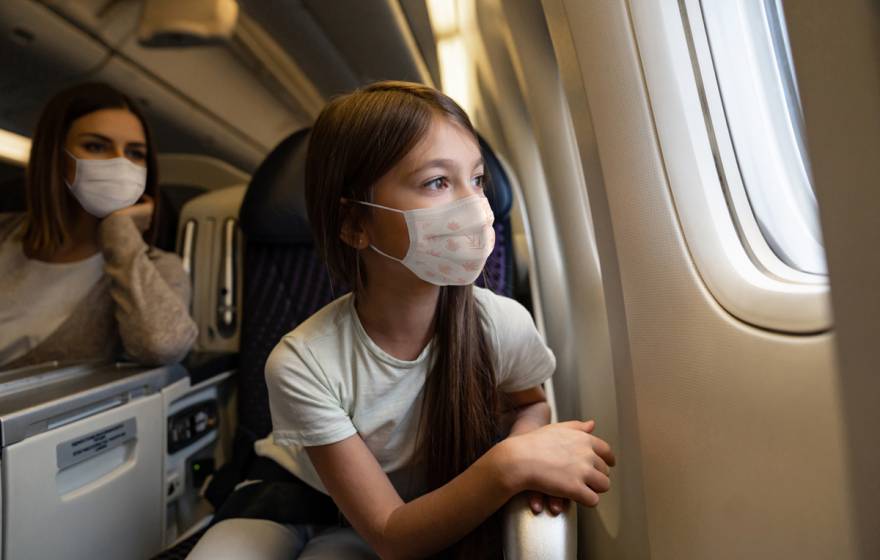 A young girl in a mask stares out an airplane window, mom in background
