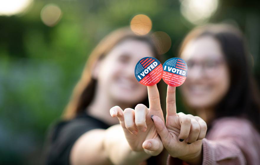 Two young women, mostly out of focus, hold up I Voted stickers to the camera, smilling