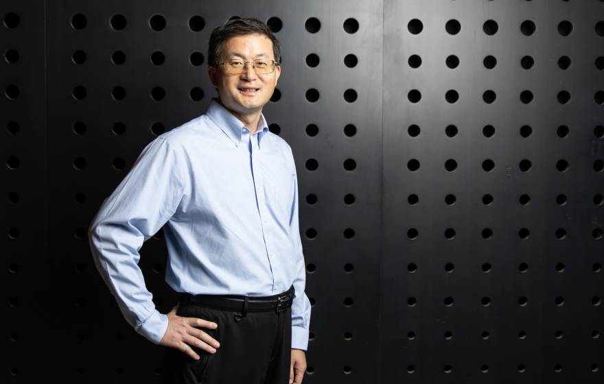 A man with glasses stands in front of a dark textured wall with his hand on his hip