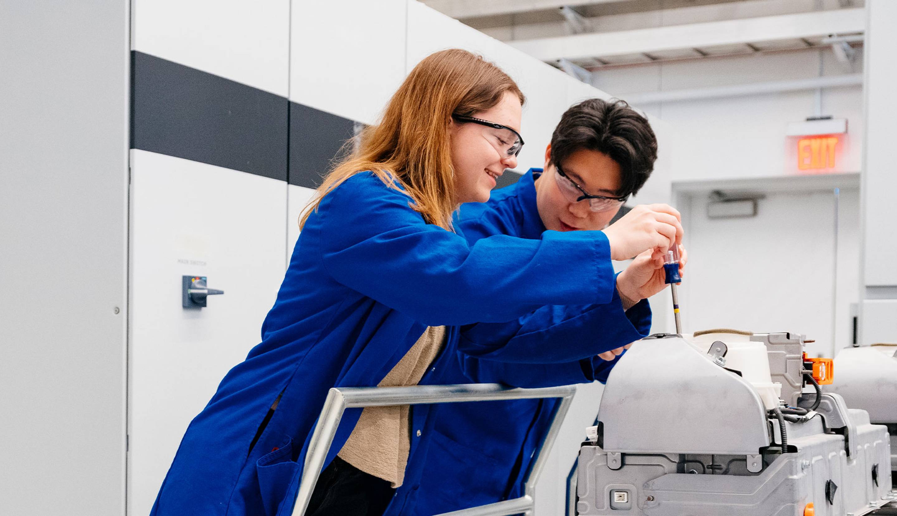 Two people in blue lab coats with goggles working on a machine with a screwdriver. 