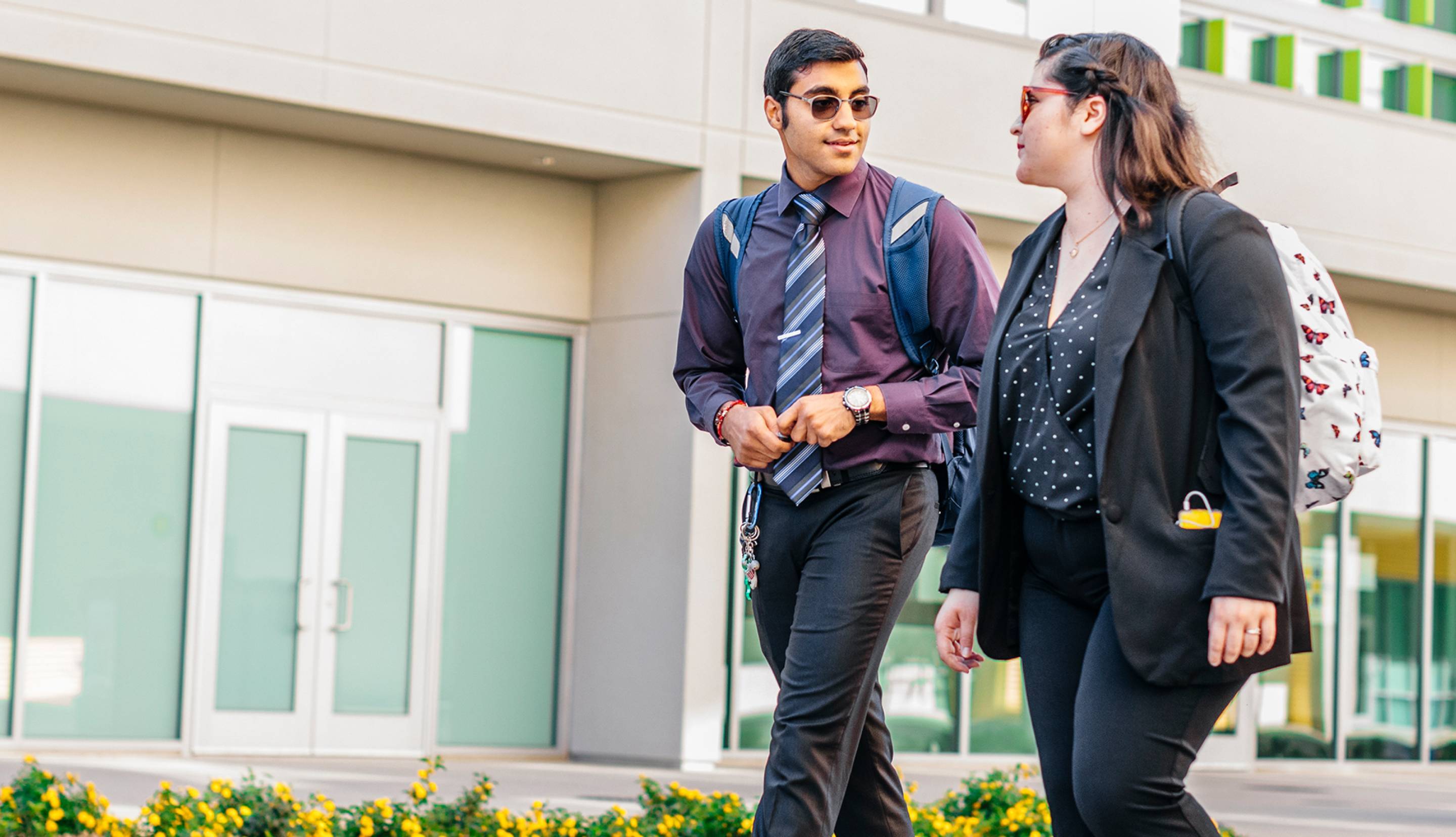 A man with a tie and a woman in business casual walk and talk outside a campus building