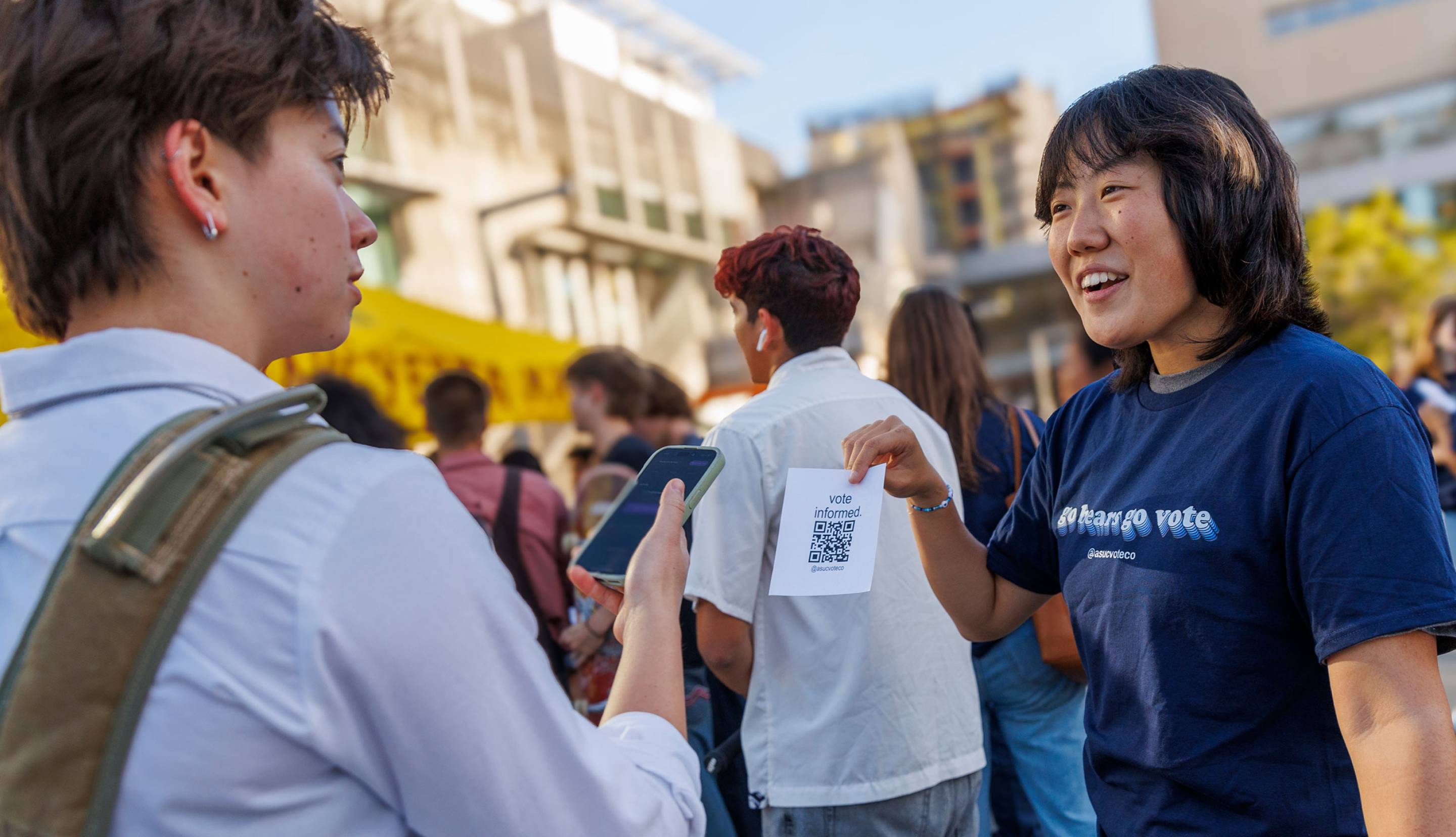 A student wearing a GO BEARS. GO VOTE. t shirt holds up a printed QR code for another student to scan with their phone