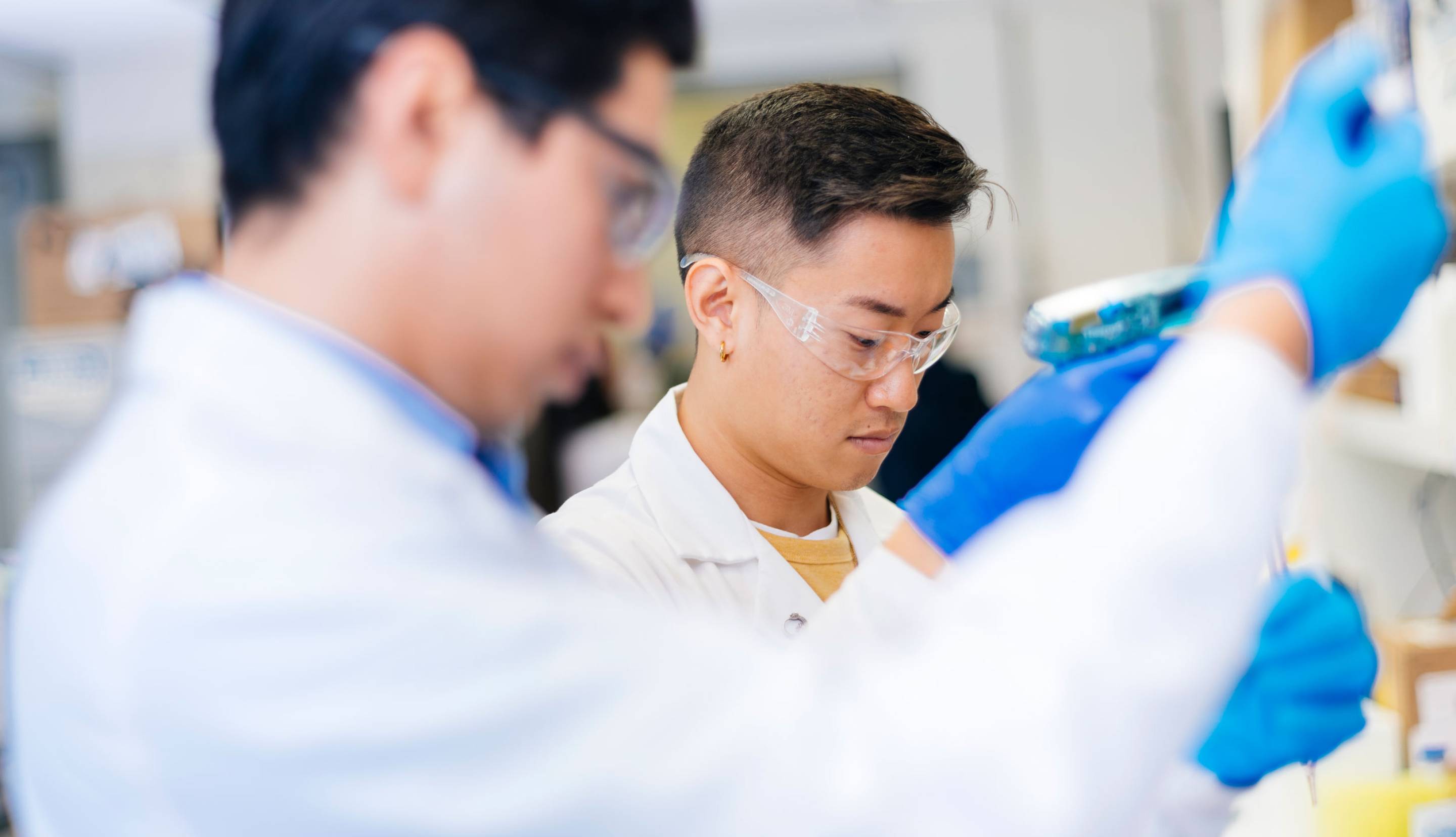 Two researchers wearing lab coats, safety glasses, and blue gloves in a lab.