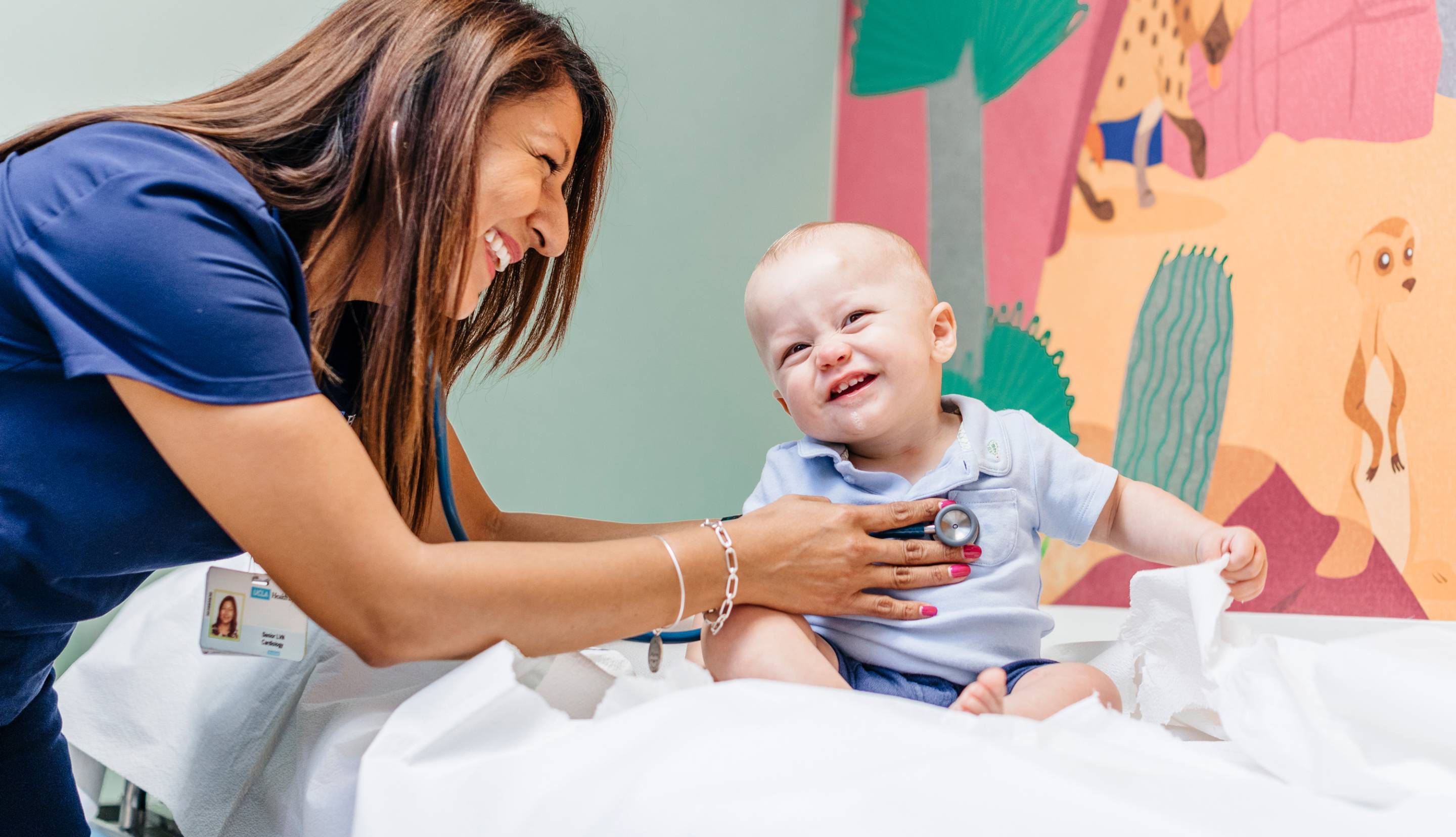 A nurse uses a stethoscope to check a baby sitting on a doctor's office table in front of a colorful mural, both with big smiles