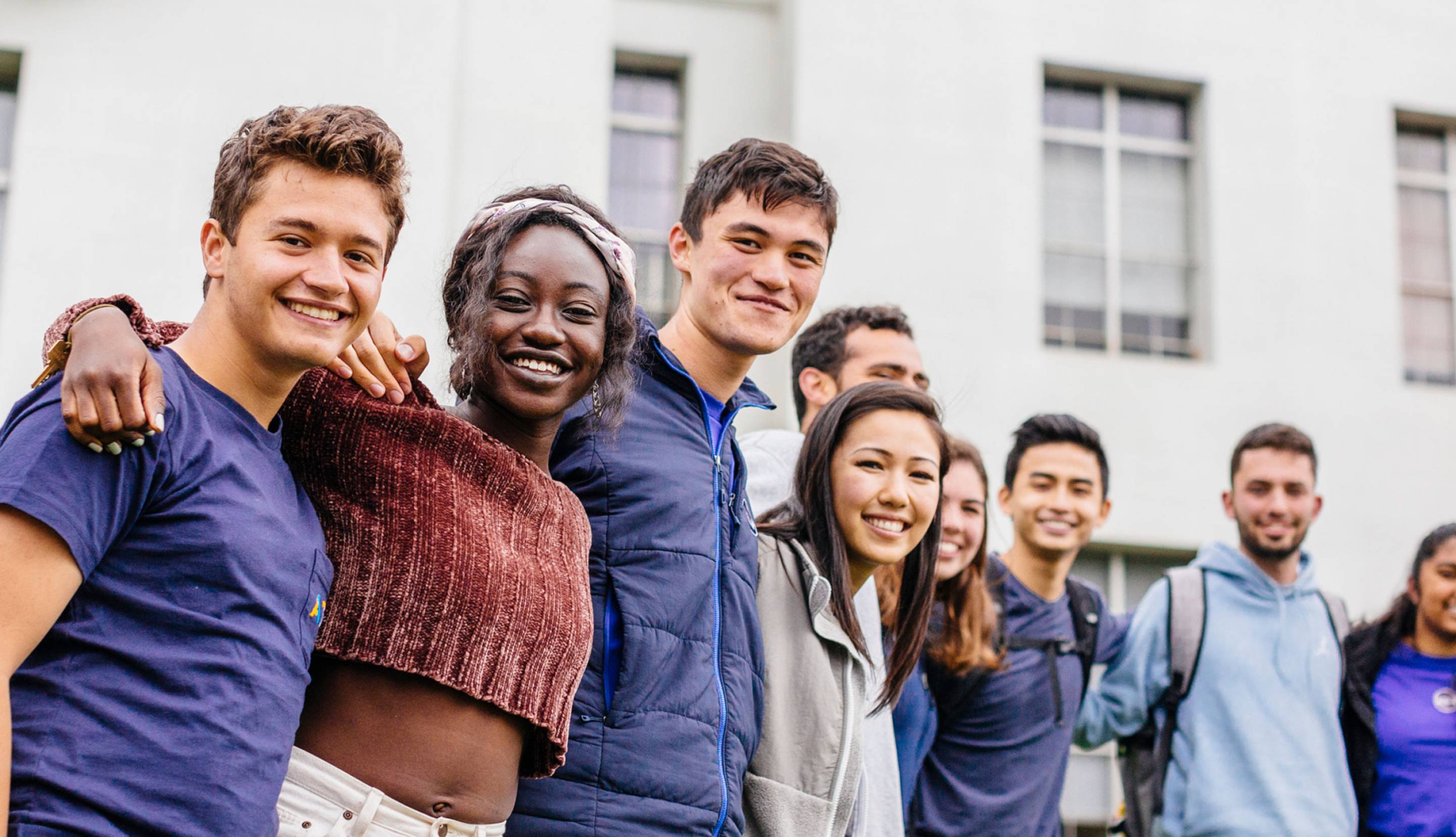 A group of students smiling with arms around each other, in front of a building with windows.