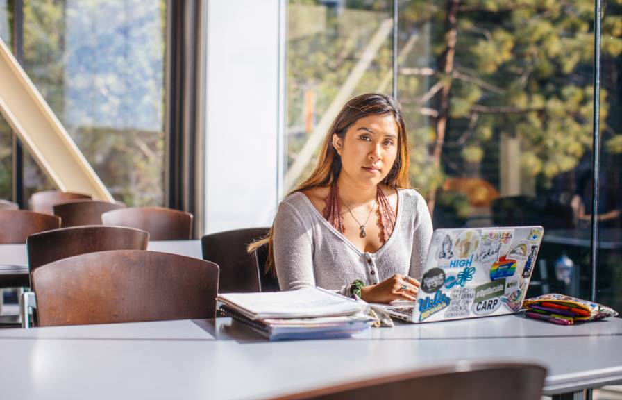 Woman at laptop in glass-paned office with lots of trees behind looks at camera