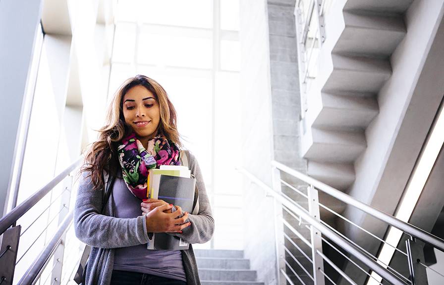 A young Latinx woman holding books smiles walking down a stairway