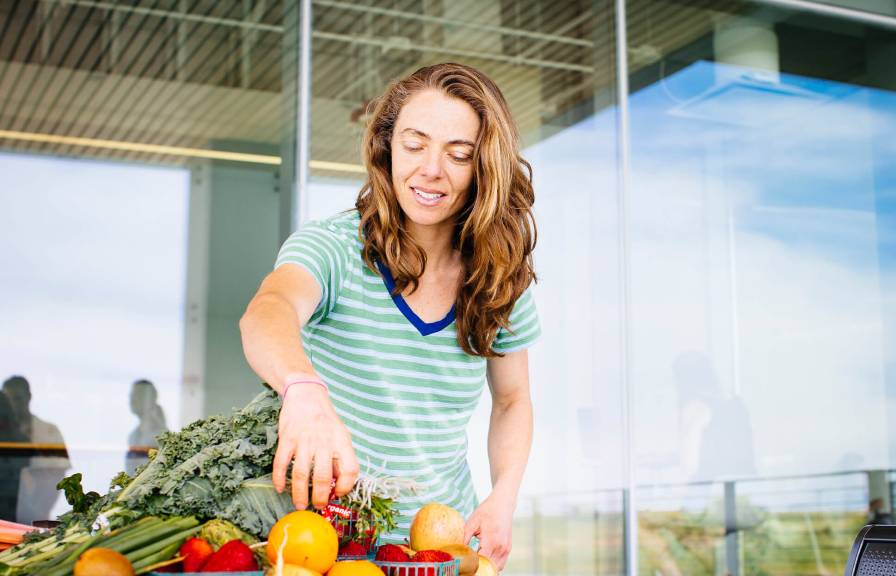 Student in front of glass building with pile of food in front picking up a vegetable. 