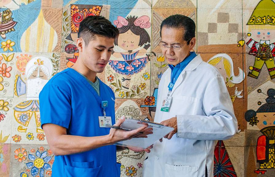 A healthcare professional in blue scrubs discusses notes on a clipboard with a doctor in a white coat.